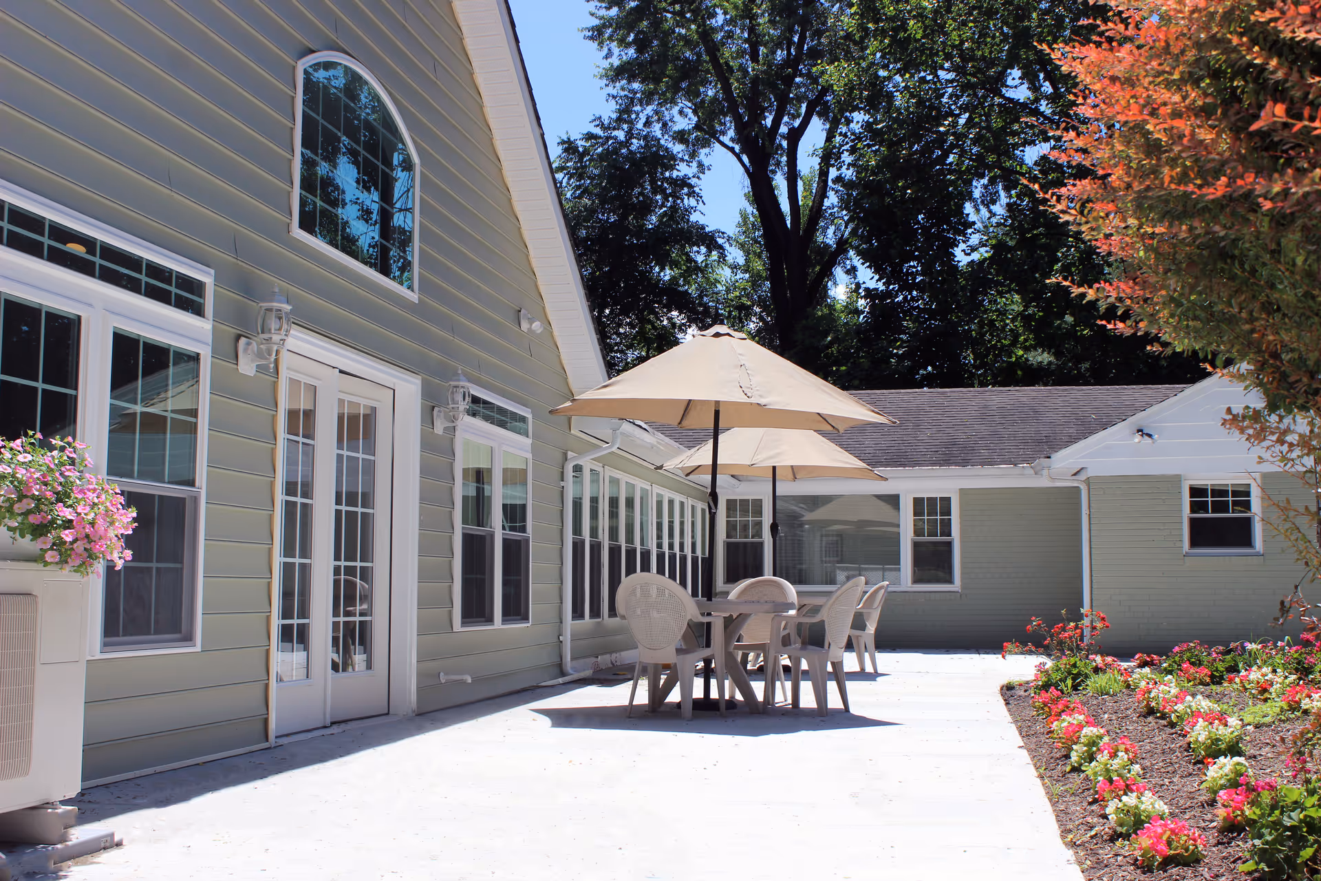 Outdoor patio area at Clifton Woods Memory Care Home with plastic tables and chairs under beige umbrellas, surrounded by a garden with colorful flowers and trees, next to a light green building with multiple windows and a glass door.