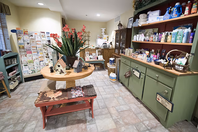 Interior view of a small shop or boutique area inside a facility, featuring a round wooden table with decorative items and flowers, a red bench underneath, green cabinets filled with various toiletries and products, and racks of greeting cards along the wall.