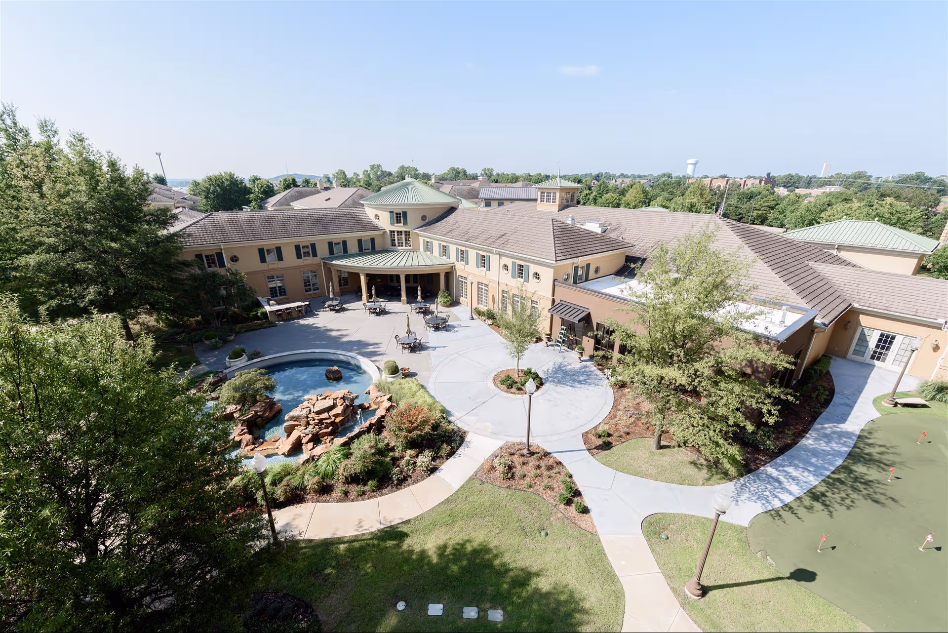 Aerial view of the Montereau senior living facility showing a large building with beige walls and green roofs surrounding a courtyard with outdoor seating, a small pond with rocks, landscaped gardens, and a putting green area. Trees and pathways are visible around the courtyard under a clear blue sky.