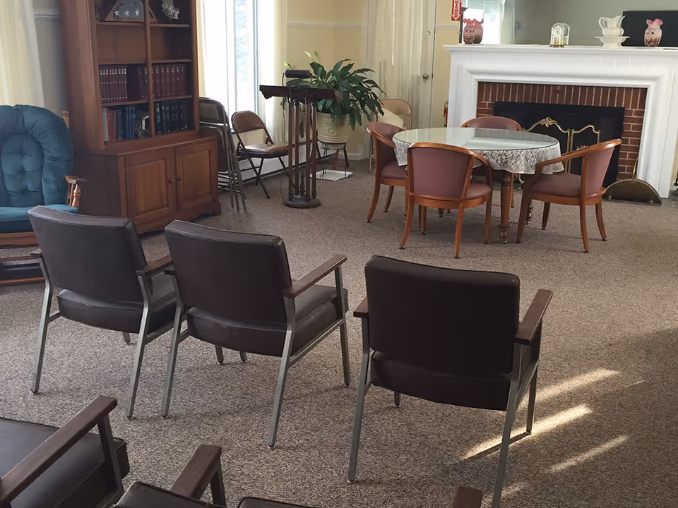 A cozy living room area with three brown chairs facing a round table covered with a lace tablecloth and surrounded by four wooden chairs. There is a fireplace with a white mantel and decorative items on top, a wooden bookshelf filled with books, a green cushioned chair, and a potted plant near a window with natural light coming in.