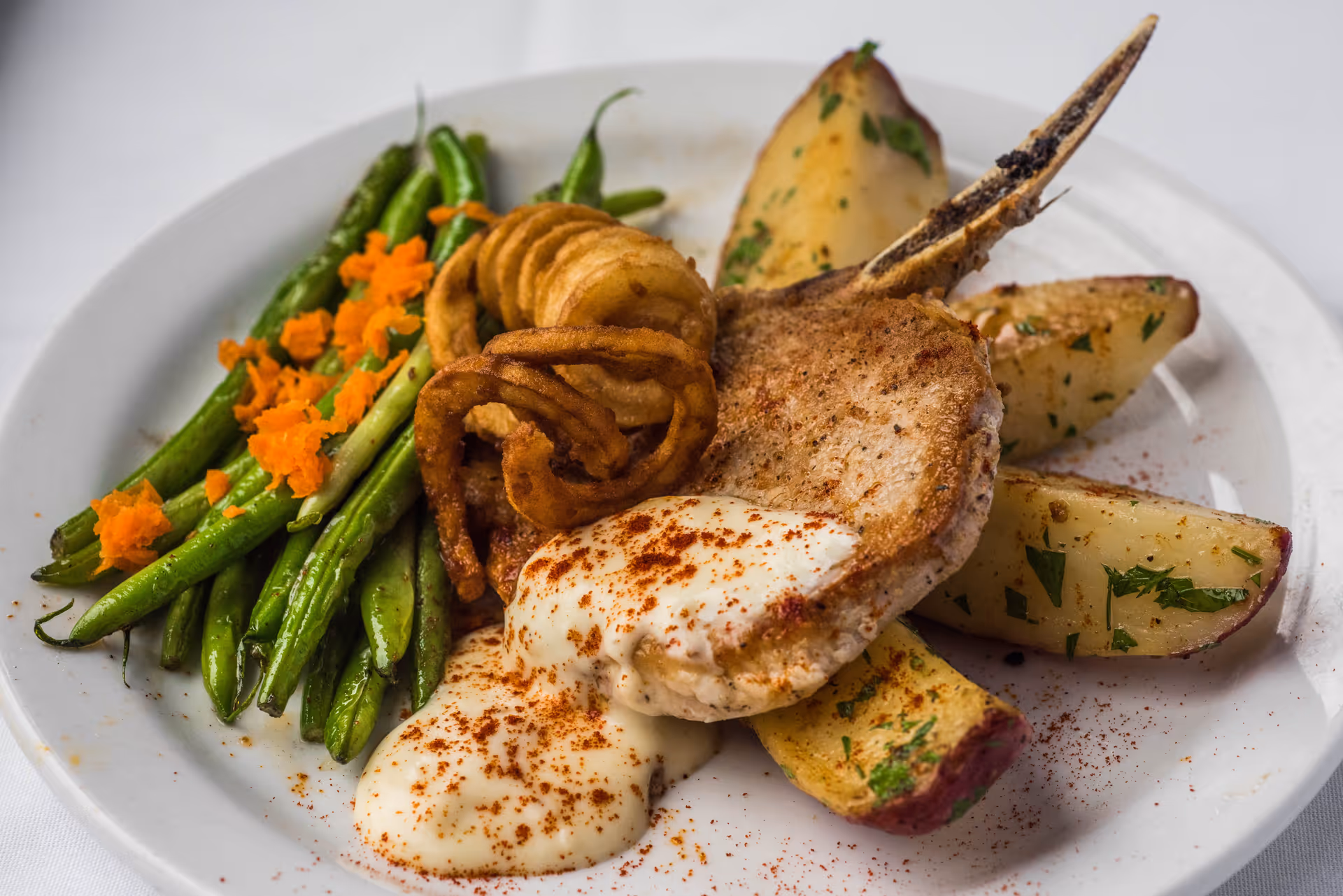 A plate of food featuring a cooked pork chop with a bone, topped with a creamy white sauce sprinkled with paprika, served with roasted potato wedges garnished with herbs, curly fried onion rings, and green beans topped with grated orange carrot.