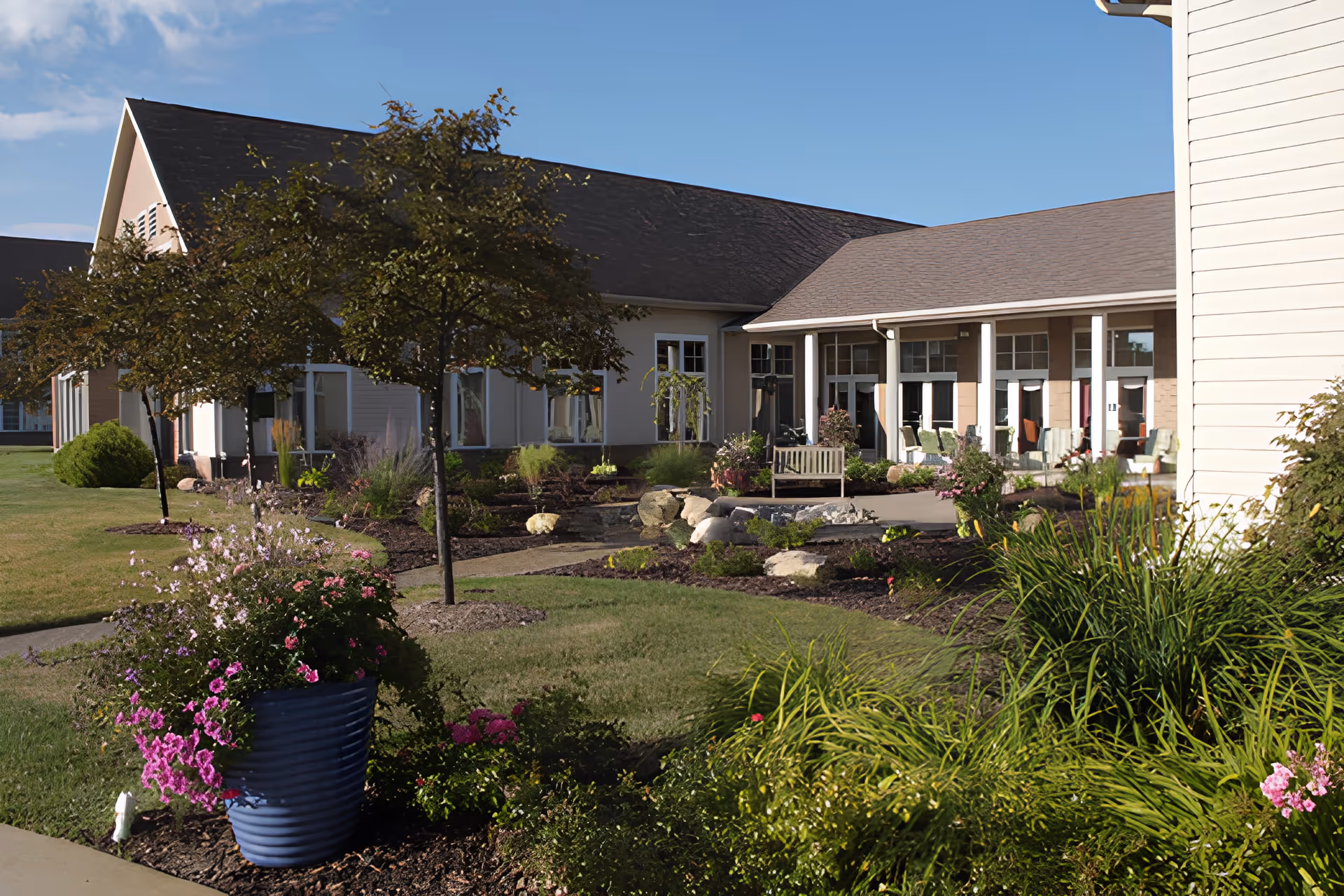 Outdoor garden area at Brecon Village featuring a well-maintained lawn, various flowering plants in a large blue pot, small trees, and a pathway leading to a building with a covered porch and seating area.