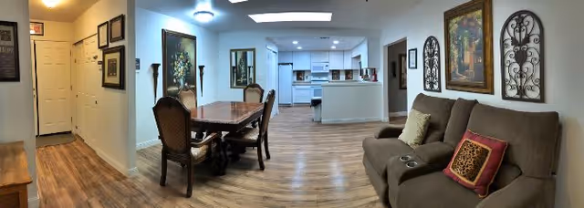 Interior view of a senior living facility showing a combined living and dining area with wood flooring. On the right, there is a brown sofa with decorative pillows and wall art above it. In the center, a wooden dining table with four chairs is positioned near the kitchen area, which features white cabinets and appliances. The left side shows a hallway with framed pictures on the walls and a door at the end.