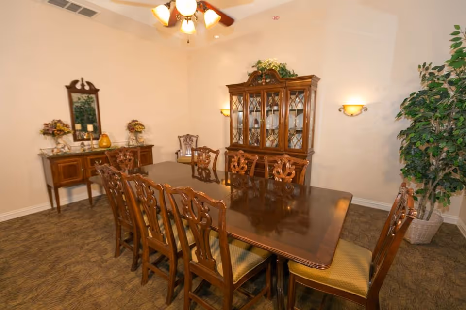 A formal dining room with a large polished wooden dining table surrounded by eight matching wooden chairs with cushioned seats. Against the back wall is a wooden china cabinet displaying plates and glassware. To the left, there is a sideboard with decorative items including flowers and candles, and a mirror hanging above it. A ceiling fan with lights is visible above the table, and a large potted plant is in the corner to the right.