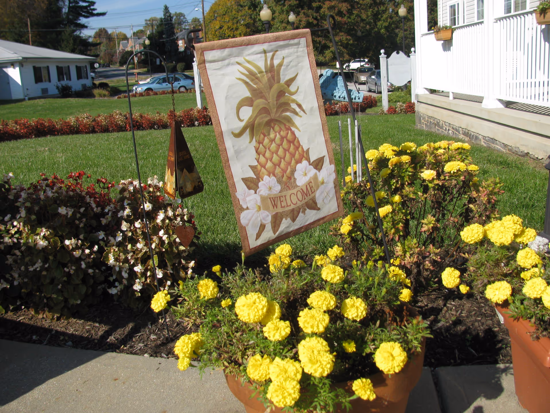 Outdoor garden area with yellow flowers in pots and flower beds, a decorative welcome flag featuring a pineapple and white flowers, a small hanging ornament, and a white porch railing on the right side of the image.