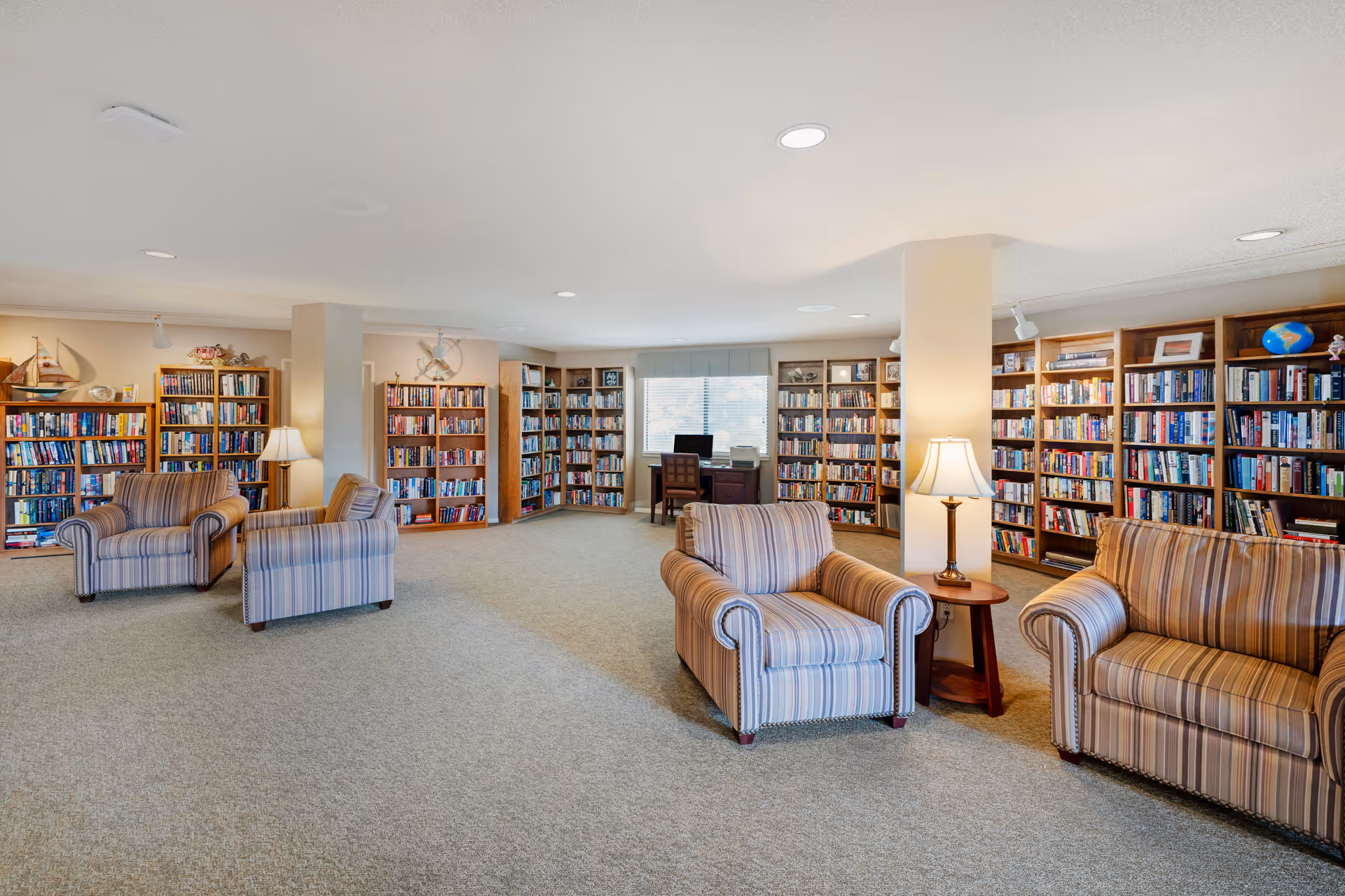 A spacious library room with multiple wooden bookshelves filled with books along the walls. There are four striped armchairs arranged in pairs with small wooden side tables and lamps between them. A desk with a computer and printer is positioned in front of a window with blinds, allowing natural light into the room. The carpeted floor and neutral-colored walls create a warm and inviting atmosphere.