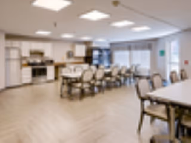 A large dining and activity room with multiple tables and chairs and a kitchenette in the background.