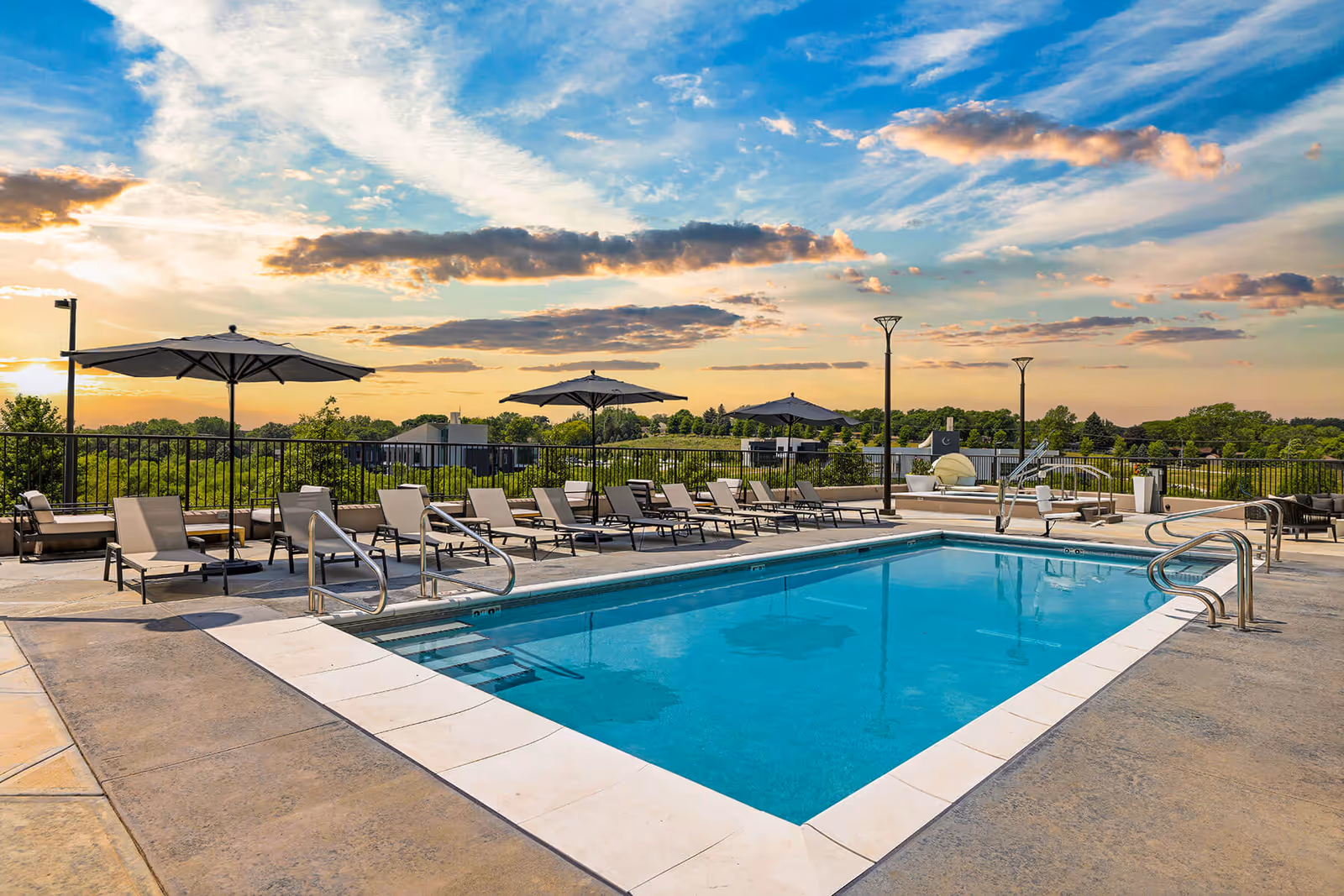Outdoor swimming pool area at sunset with multiple lounge chairs and umbrellas arranged around the pool, surrounded by a black metal fence and trees in the background.