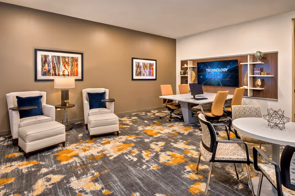 A modern meeting or lounge room with two beige armchairs with dark blue pillows and a small round table between them on the left. The room has a patterned carpet with orange, gray, and white colors. On the right side, there is a white table with four orange office chairs and a laptop on it. Behind the table is a wall-mounted TV screen displaying 'TECHNOLOGY NEWS' with shelves holding decorative items. In the foreground, there is a round white table with four patterned chairs around it and a geometric decorative object on top.