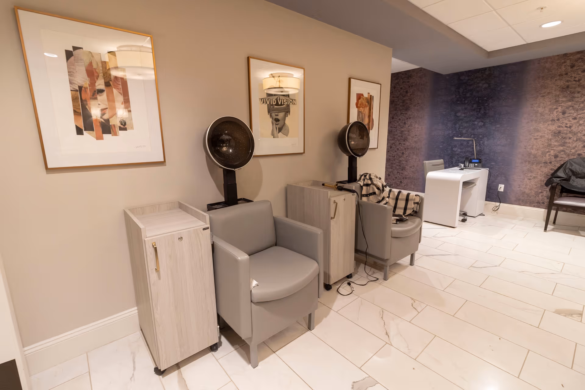 Interior view of a salon area in a senior living facility with two gray salon chairs under hair dryers, two light wood cabinets, framed artwork on the beige wall, a white desk with a chair and phone, and a dark textured accent wall in the background.