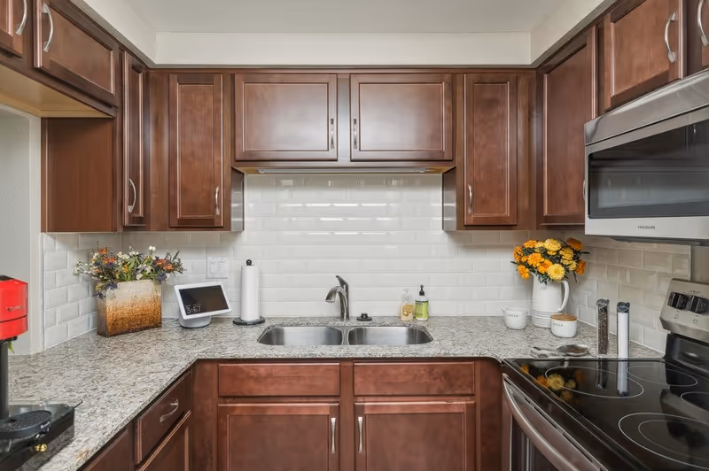 A modern kitchen with dark wooden cabinets, a granite countertop, a double stainless steel sink, and a white subway tile backsplash. On the left side of the counter, there is a red coffee maker, a decorative flower arrangement, a digital clock, and a paper towel holder. On the right side, there is a white pitcher with yellow flowers, two small white containers labeled 'cream' and 'sugar', and salt and pepper grinders. A stainless steel microwave and electric stove are also visible.