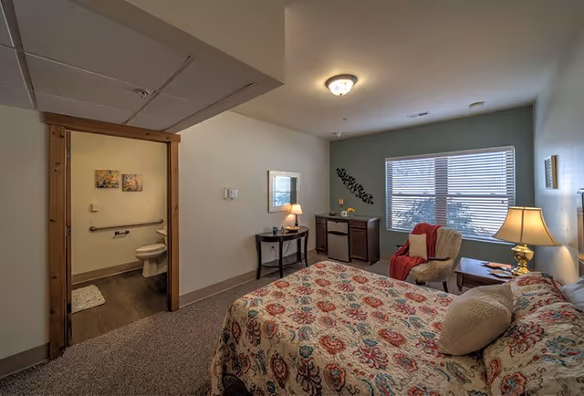 A cozy senior living bedroom with a patterned bedspread, a bedside table with a lamp, an armchair with a red throw, a small table with a lamp and mirror, and a window with blinds. An open door reveals an adjacent bathroom with a toilet and grab bars.