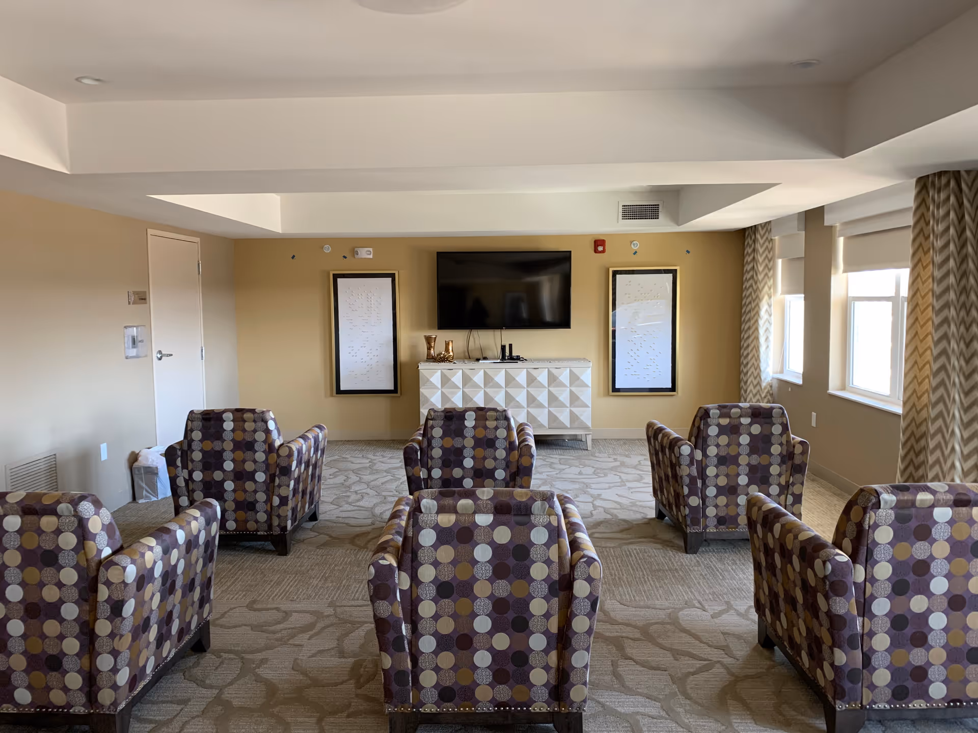 A common area with six patterned armchairs arranged in two rows facing a wall-mounted flat screen TV. Below the TV is a white geometric cabinet with decorative items on top. The room has beige walls, carpeted floor with a floral pattern, and windows with chevron-patterned curtains on the right side.