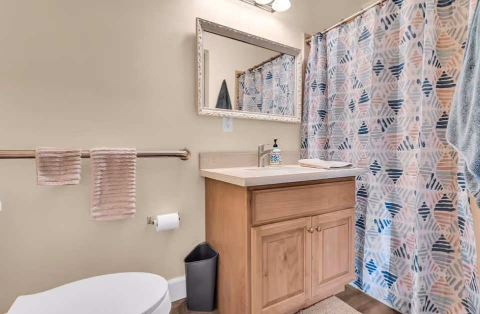 A bathroom with a wooden vanity cabinet topped with a white sink and a soap dispenser. Above the sink is a rectangular mirror with a decorative frame. To the left, there is a toilet with a toilet paper holder on the wall and two beige hand towels hanging on a metal towel bar. On the right side, a shower curtain with a geometric pattern in shades of blue, pink, and white is visible. A small black trash bin is placed on the floor next to the vanity.