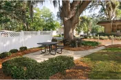 Outdoor seating area with a picnic table on a concrete path surrounded by greenery, bushes, and large trees. A white fence and a building are visible in the background.