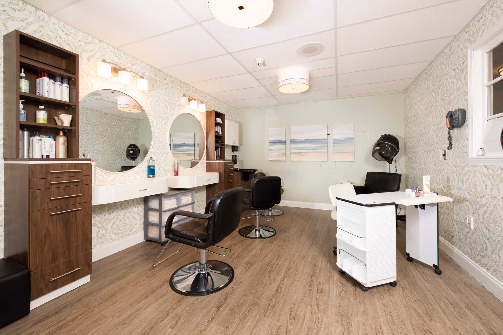 Interior of a senior living facility salon with two black salon chairs in front of two round mirrors mounted on a wall with patterned wallpaper. There are shelves with hair care products, a hair dryer chair, and a manicure table with nail polish. The floor is wood, and there are three abstract paintings on the far wall.