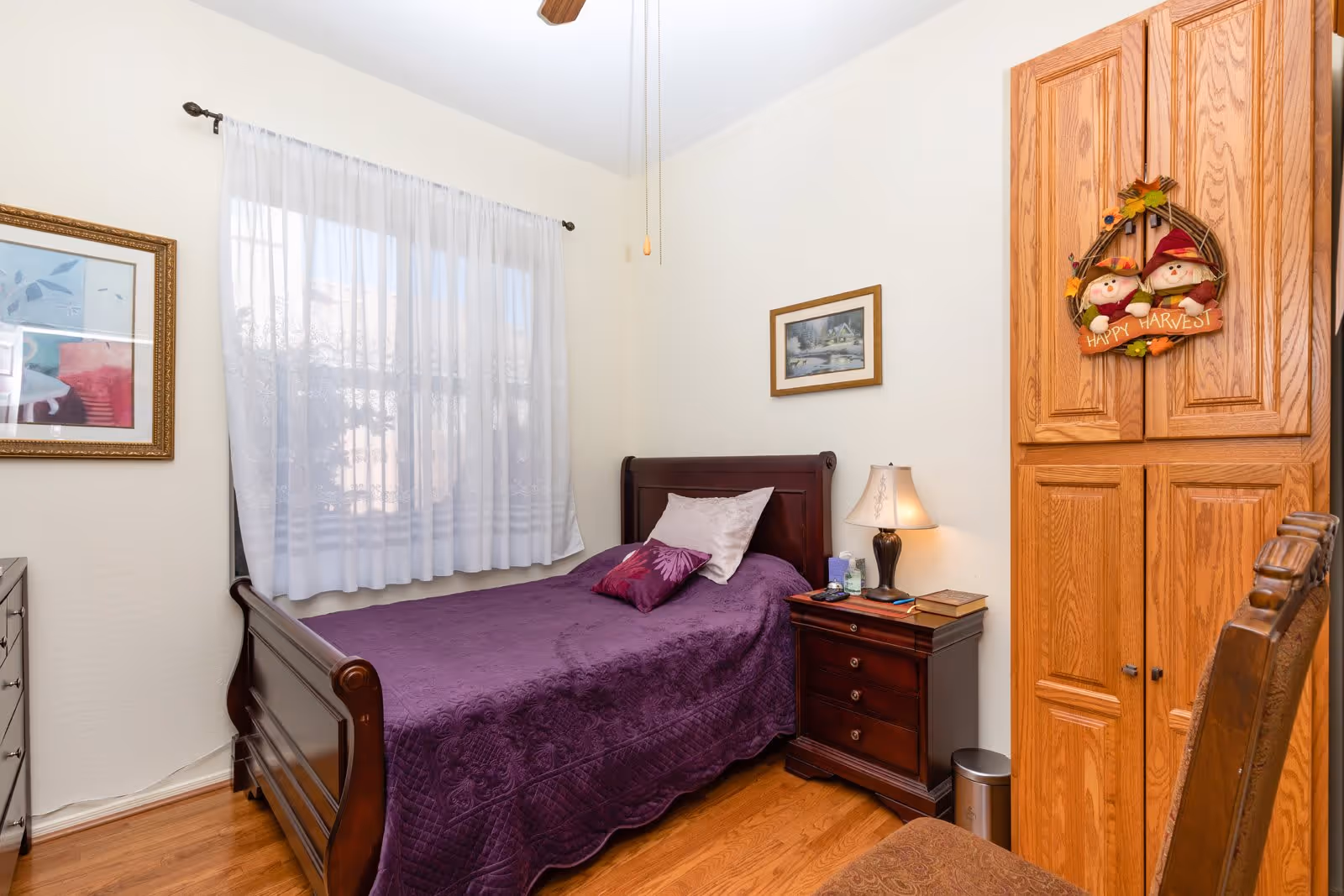A small bedroom with a single dark wooden bed covered with a purple quilt and two pillows. Next to the bed is a dark wooden nightstand with a lamp, books, and some small items. There is a wooden wardrobe with a decorative 'Happy Harvest' wreath hanging on it. A window with sheer white curtains lets in natural light, and framed pictures hang on the walls. The floor is wooden, and part of a wooden chair is visible in the foreground.
