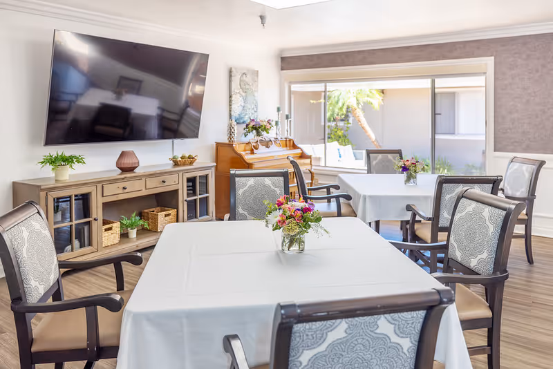 A bright and clean dining room with tables covered in white tablecloths, each adorned with a small vase of colorful flowers. The room features comfortable chairs with patterned upholstery, a large flat-screen TV mounted on the wall, a wooden sideboard with decorative items, and a piano near a large window letting in natural light.