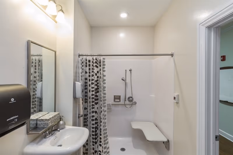A clean, accessible bathroom with a white sink, a wall-mounted mirror, a soap dispenser, a paper towel dispenser, and a shower area with a grab bar, handheld showerhead, and a fold-down shower seat. The shower curtain has a pattern of large black dots on a white background.