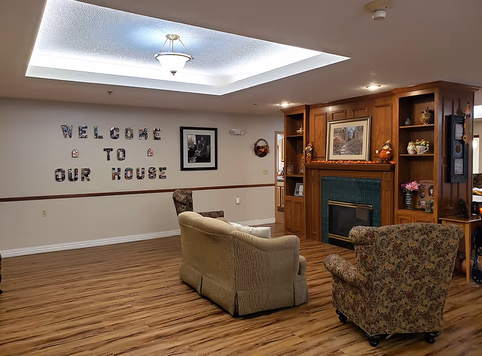 A cozy senior living common room with sofas and armchairs facing a fireplace and a wall sign reading "WELCOME TO OUR HOUSE".
