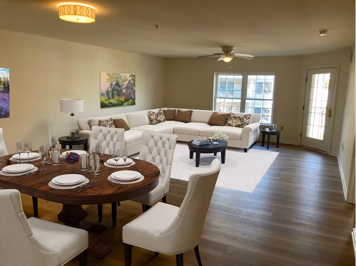 A bright and spacious living and dining area in a retirement community apartment. The room features a large beige sectional sofa with decorative pillows, a round wooden dining table set with plates, glasses, and cutlery, and several upholstered dining chairs. There are two paintings on the wall, a ceiling light fixture, a ceiling fan, and large windows letting in natural light. The floor is wooden with a light-colored rug under the sofa area.