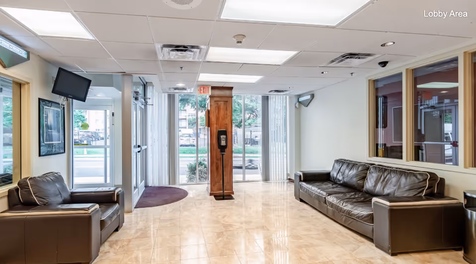 A bright and clean lobby area with large windows and glass doors letting in natural light. The space features two dark brown leather sofas facing each other, a wooden stand with a hand sanitizer dispenser in the center, and a wall-mounted TV on the left side. The floor is tiled with a light beige color, and there are vertical blinds on the windows. An exit sign is visible above the door, and the words 'Lobby Area' are displayed in the top right corner.
