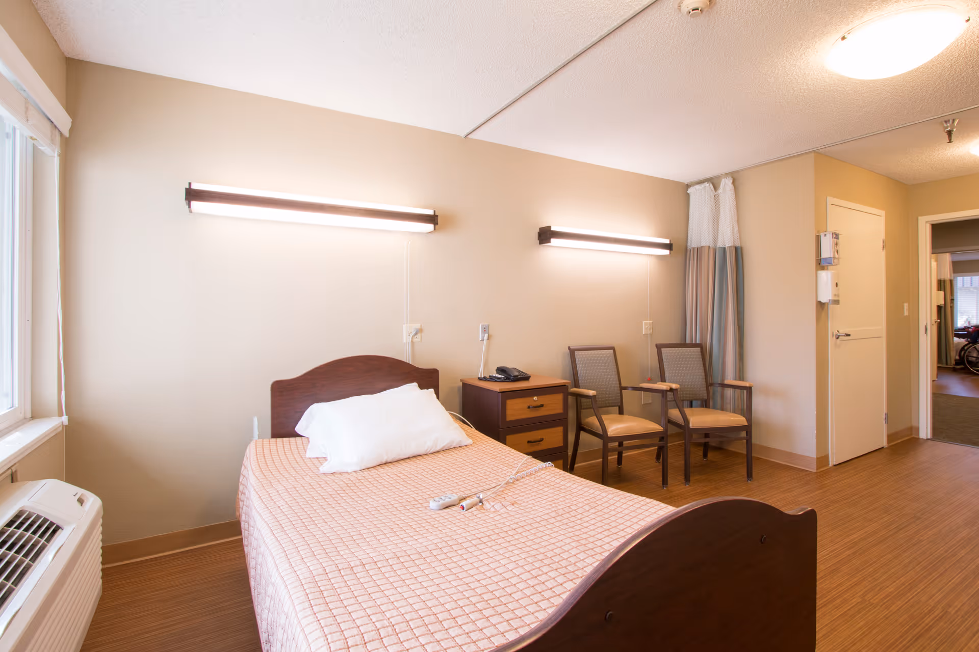 A simple, clean bedroom in a senior living facility with a single bed covered in a pink checkered blanket and a white pillow. Two wall-mounted lights illuminate the room above the bed. Next to the bed is a small wooden nightstand with a telephone on it. Two wooden chairs with cushioned seats are placed against the wall near a curtain. A window with blinds and an air conditioning unit are visible on the left side. The room has light-colored walls and wood flooring, with a door leading to another area in the background.