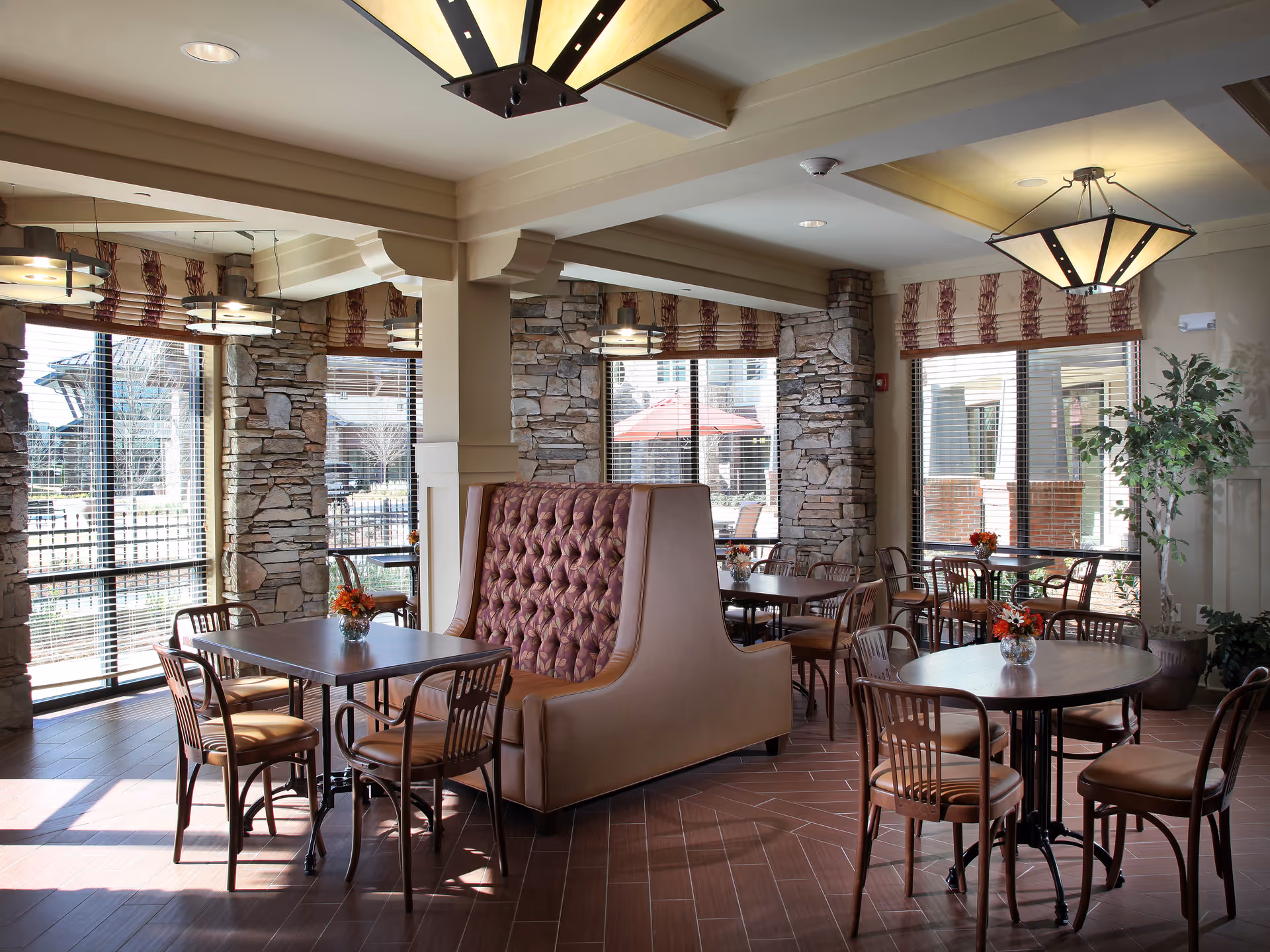 A bright dining area with multiple tables and chairs, featuring a large tufted booth in the center. The room has stone accent walls, large windows with blinds, and decorative light fixtures hanging from the ceiling. Small floral arrangements are placed on each table.