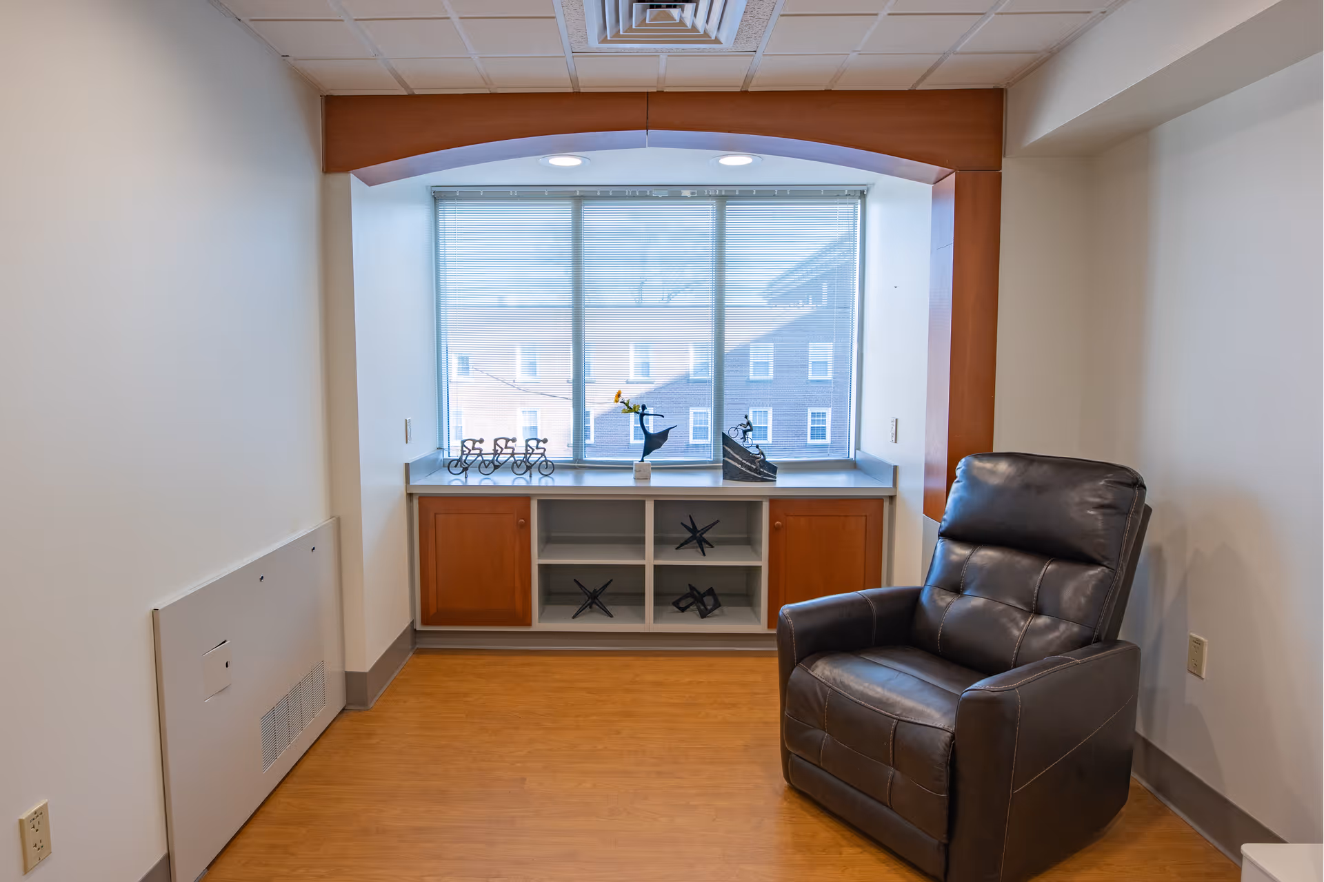 A small room with a large window featuring blinds, a built-in cabinet with wooden doors and open shelves displaying decorative items, and a dark brown leather recliner chair positioned to the right. The room has light-colored walls, a wooden floor, and a ceiling with recessed lighting.