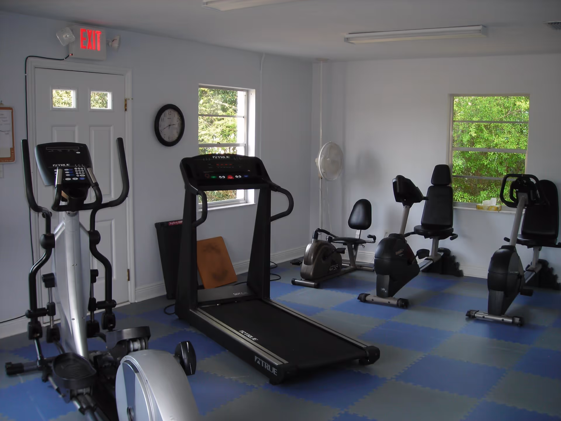 A small fitness room with exercise equipment including a treadmill, elliptical machine, and two recumbent bikes. The room has blue and gray interlocking foam floor tiles, white walls, two windows showing green foliage outside, a wall clock, a standing fan, and an exit door with a red exit sign above it.