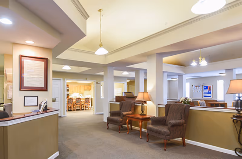 Reception and common seating area in a senior living facility with armchairs, side tables, lamps, and a dining area visible in the background.