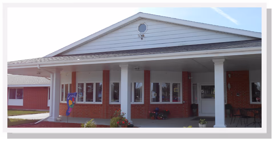 Front exterior view of a single-story brick building with white columns supporting a covered porch. There are several windows and a door visible, along with some outdoor seating and potted plants. A colorful welcome flag is displayed near the entrance.