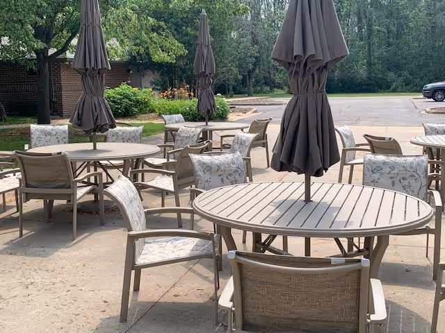 Outdoor patio area with multiple round tables and chairs arranged around them. Each table has a closed umbrella in the center. The patio is surrounded by trees and greenery, with a parking lot visible in the background.