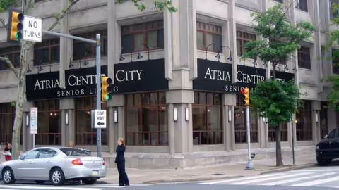 Corner of Atria Center City Senior Living building with street, cars, and a pedestrian.