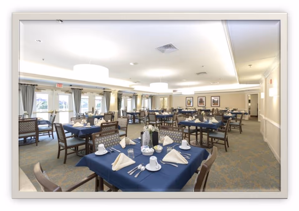 A spacious dining room with multiple tables covered in blue tablecloths, each set with white napkins, cups, glasses, and silverware. The room has large windows with gray curtains, soft overhead lighting, and framed artwork on the walls.