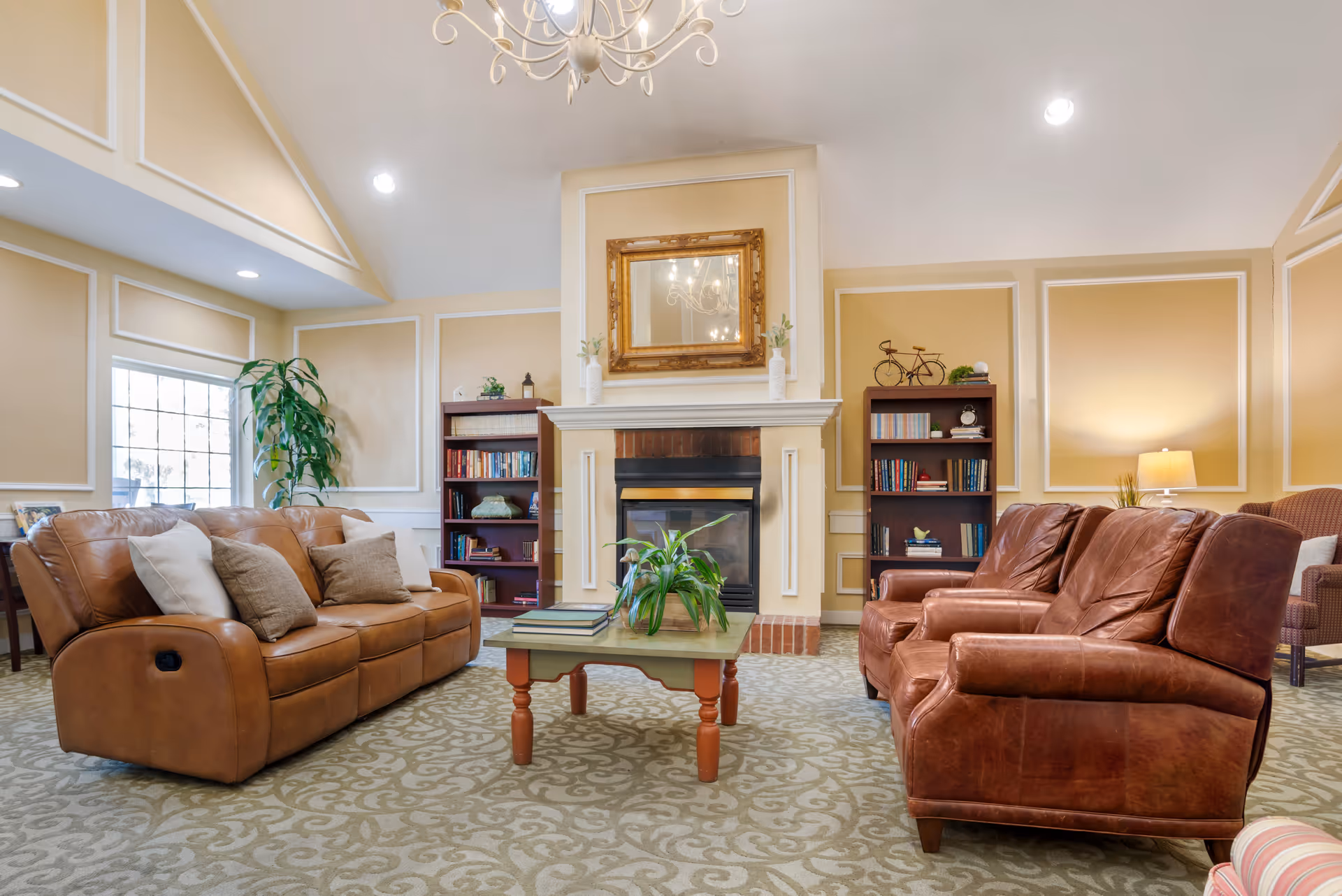 A cozy living room with beige walls and white trim, featuring a brown leather sofa and two matching leather armchairs arranged around a wooden coffee table with books and a plant. There is a fireplace with a gold-framed mirror above it, flanked by two bookshelves filled with books and decorative items. A large window lets in natural light, and a chandelier hangs from the ceiling.