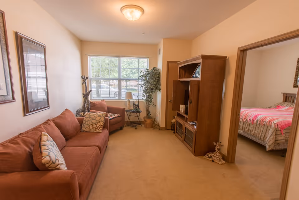A cozy living room with a brown sofa and matching armchair, decorated with patterned cushions. There is a wooden entertainment center on the right side and a window with blinds letting in natural light. A potted plant and small decorative giraffe statues are also visible. An open doorway leads to a bedroom with a bed covered in a pink and beige quilt.
