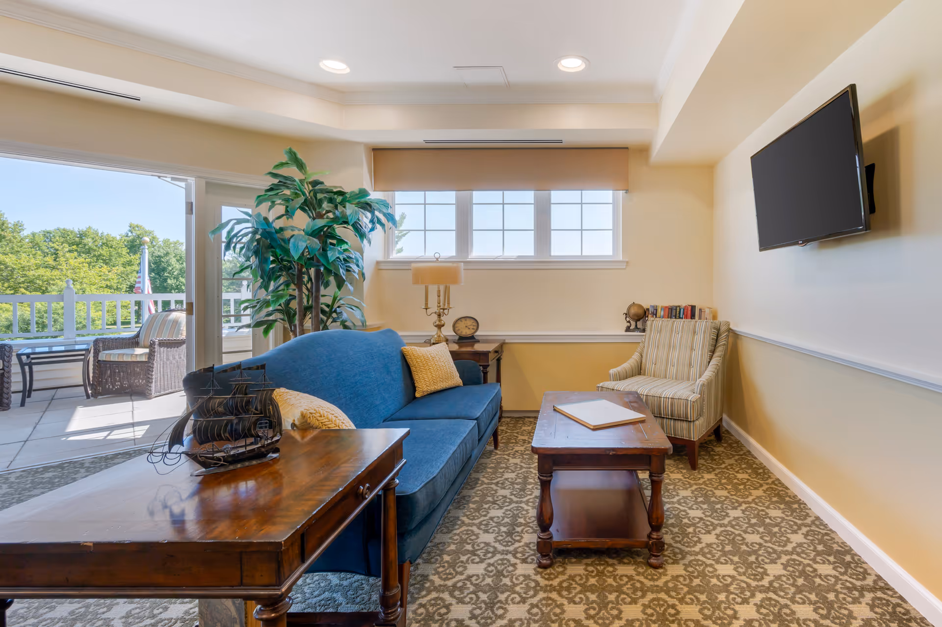 Bright common living room with a blue sofa, armchair, wooden coffee tables, wall-mounted TV and sliding doors opening to an outdoor patio.