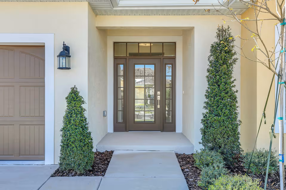Front entrance of a building with a brown door featuring glass panels and sidelights, flanked by two tall green shrubs. There is a beige wall with a black outdoor lantern on the left side and a garage door partially visible on the left.