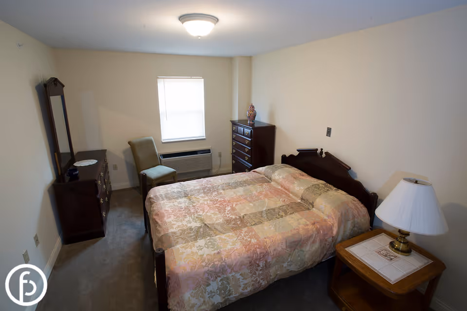 Small furnished bedroom with a double bed, nightstand and lamp, dresser and chair under a window.