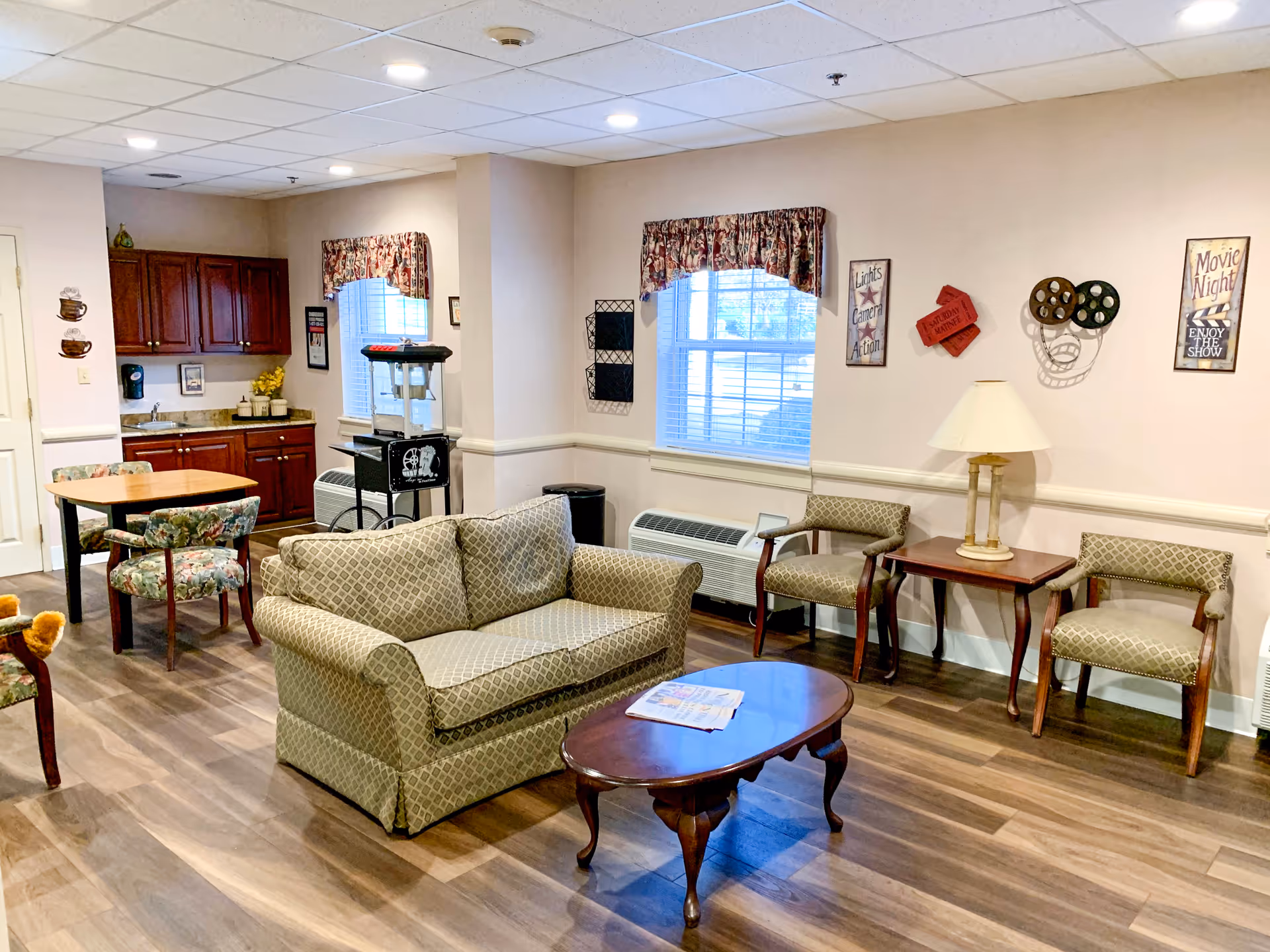 A cozy common area in a senior living facility featuring a patterned loveseat and two matching armchairs around a wooden coffee table with a newspaper on it. There is a small table with two floral upholstered chairs near a kitchenette with wooden cabinets and a popcorn machine. The walls are decorated with movie-themed signs and a lamp on a side table. The room has wood flooring and two windows with floral valances.