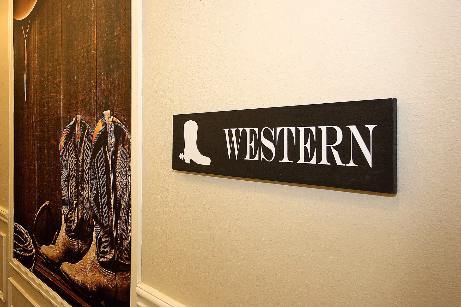 A hallway wall with a black sign that has a white cowboy boot icon and the word 'WESTERN' written on it. Next to the sign is a large picture of cowboy boots hanging on a wooden wall.
