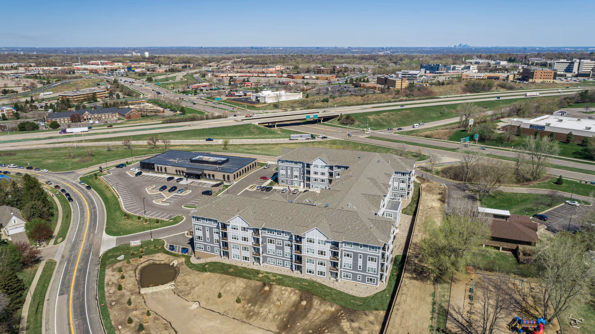 Aerial view of a large senior living facility building with multiple floors, surrounded by parking lots, roads, and green spaces. The facility is located near a highway with visible traffic and other commercial buildings in the distance under a clear blue sky.