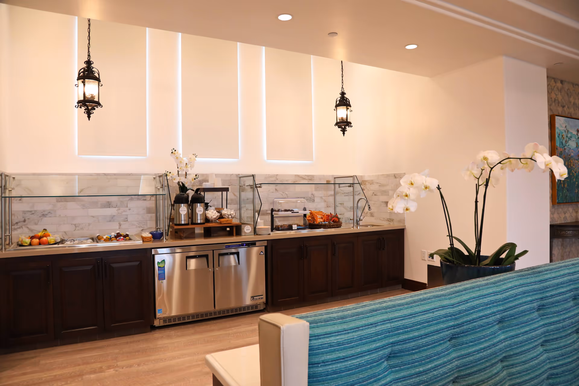 A modern kitchenette area with dark wooden cabinets, a stainless steel refrigerator, and a countertop with a sink. The countertop holds coffee dispensers, snacks, plates, and a fruit basket. Two decorative hanging lantern lights are suspended from the ceiling. A blue-striped couch and a potted orchid plant are visible in the foreground.