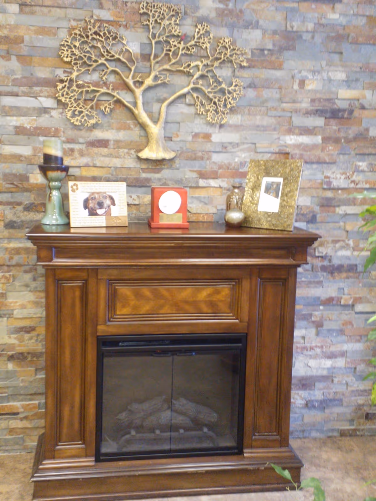 A wooden electric fireplace with a glass front set against a stone brick wall. On top of the fireplace mantel are decorative items including a green candle holder with a candle, a framed photo of a dog, a red award plaque, a small decorative bottle, a metallic egg-shaped object, and a gold picture frame with a photo. Above the mantel is a large decorative metal wall art piece shaped like a tree.
