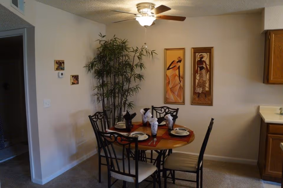 A dining area with a round wooden table set for four with plates, napkins, and utensils. Four black metal chairs with light-colored cushions surround the table. There is a tall green plant in the corner, two framed artworks on the wall, a ceiling fan with a light above, and part of a kitchen counter with wooden cabinets visible on the right.