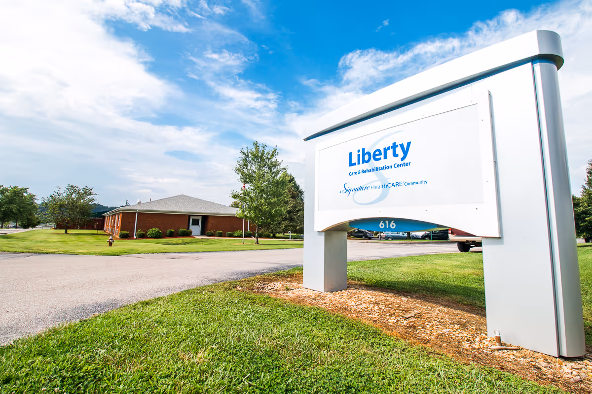 Outdoor view of Liberty Care and Rehabilitation Center sign with a building and parking lot in the background under a partly cloudy blue sky.