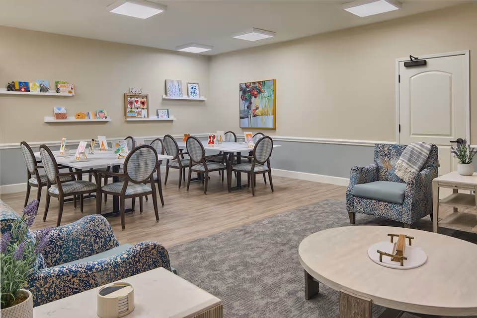 A well-lit room with a dining area featuring two tables surrounded by chairs, and a seating area with patterned armchairs and a round coffee table. The walls are decorated with shelves holding small art pieces and a colorful painting. The floor is a combination of wood and carpet.