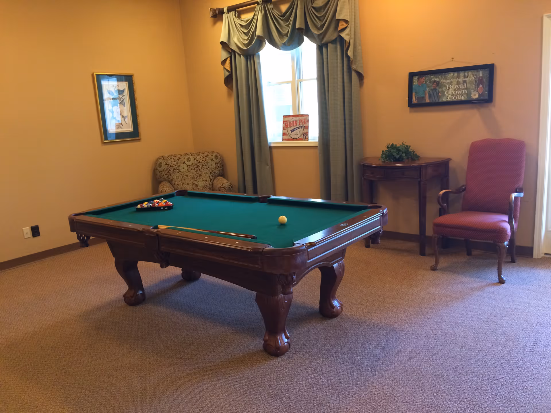 Carpeted common room with a green-felt pool table, upholstered chairs, a small side table, and a window with drapes.