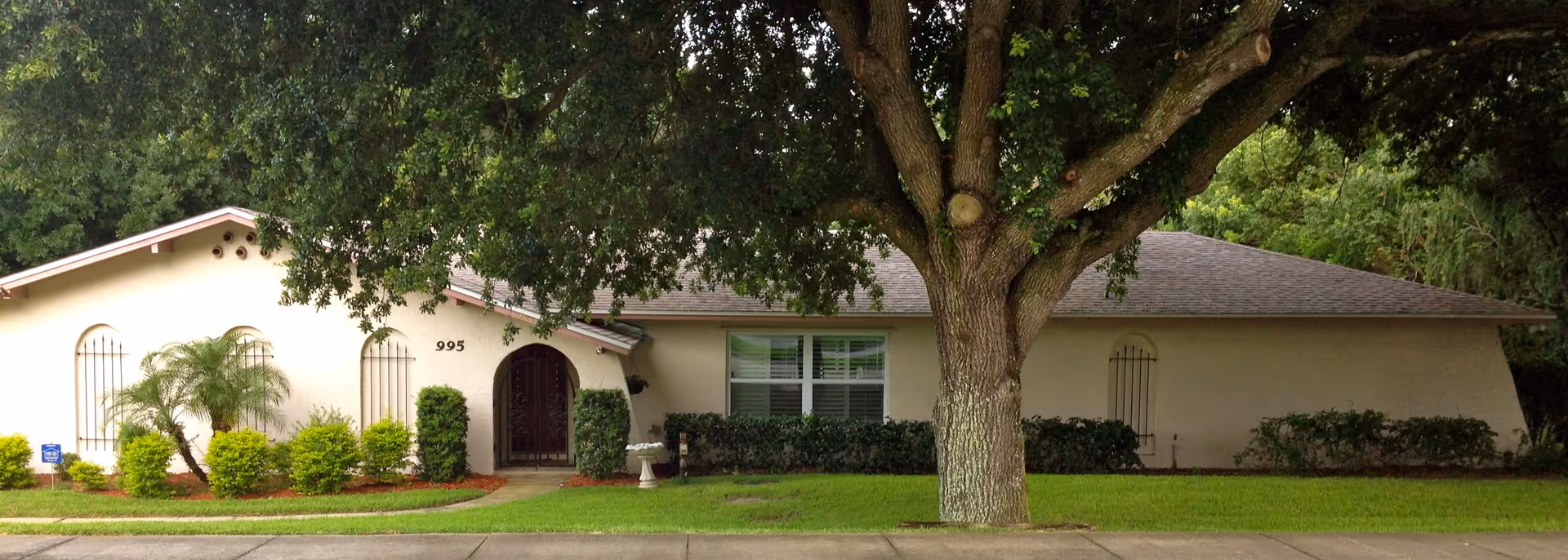 Single-story residential building with a light-colored exterior, a large tree in the front yard, green shrubs along the front, and a sidewalk leading to a gated front door. The house number 995 is visible near the entrance.